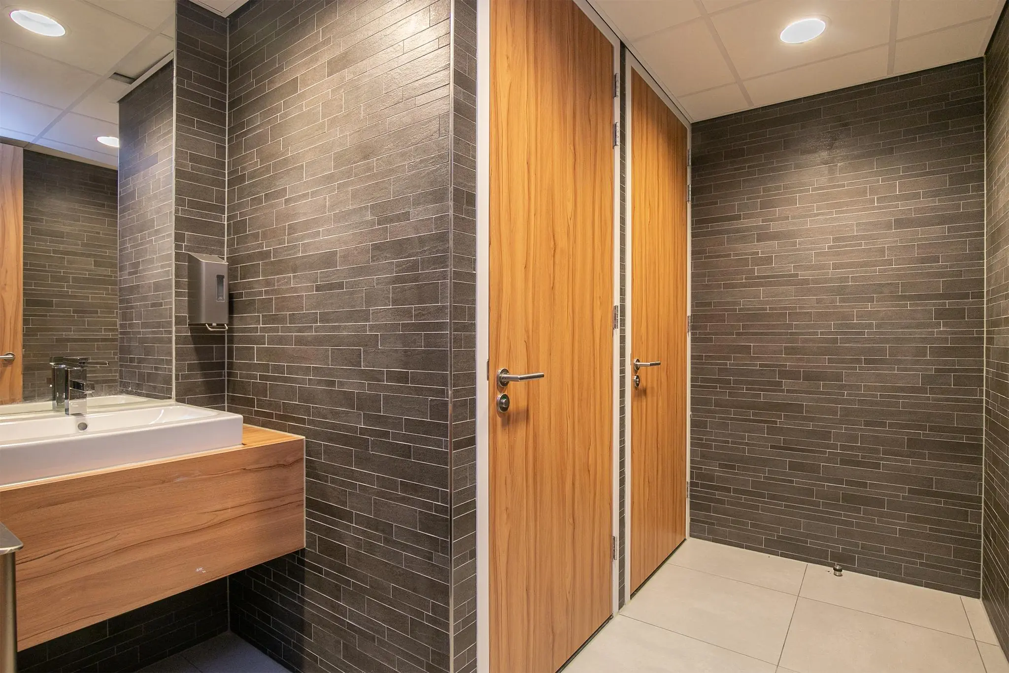 Moderne toiletruimte in Rivium Quadrant met houten deuren, donkergrijze wandtegels en een wastafel met houten meubel.